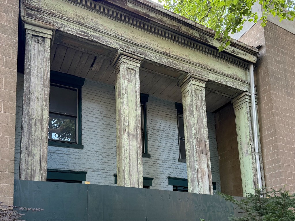 Close-up of a weathered, two-story building featuring four large, peeling columns, with green accents and a backdrop of a brick wall.