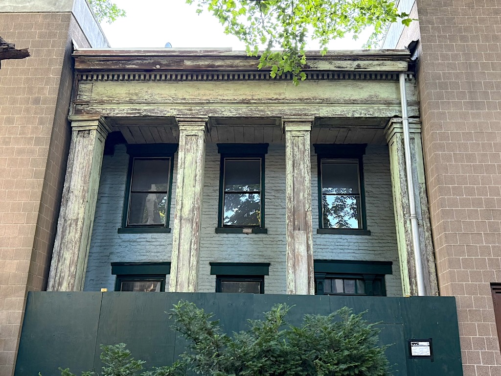 Front view of a building with three worn columns and windows, surrounded by modern structures and greenery.