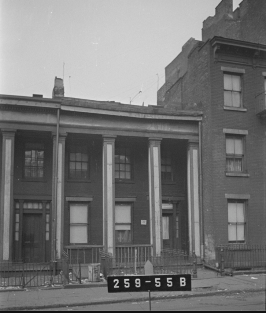 Black and white image of a row of two-story brick buildings with columns, showing a partially visible address sign, and a street view featuring fences and debris.