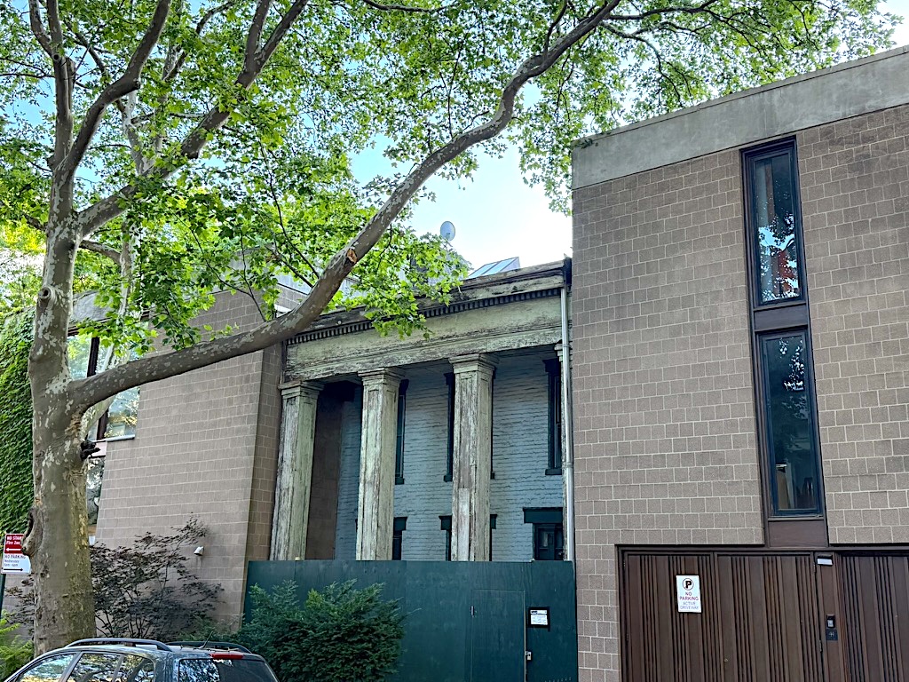 A partially visible building with classical columns set against a modern brick structure, surrounded by green trees and foliage.