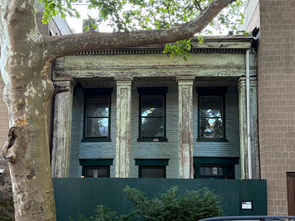 Front view of an old building with tall, greenish columns and large windows, partially obscured by a tree and surrounded by modern structures.