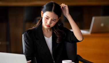 A woman in business attire sits at a desk with a laptop, looking at the screen with a confused expression while scratching her head. An open notebook, coffee cup, and another laptop are visible in the background.