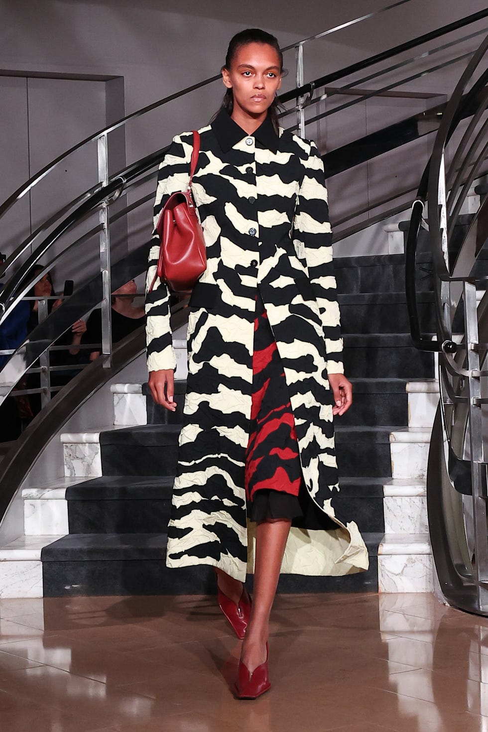 Model walking down stairs in a black and white patterned coat, red dress, and carrying a red handbag.