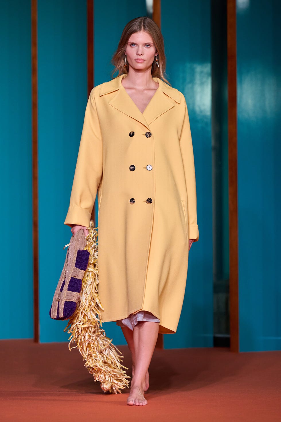 Model wearing a yellow coat and holding a textured handbag, walking on a runway featuring a turquoise backdrop.