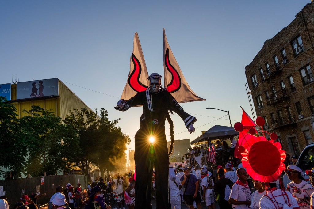 A person on stilts in a sailor costume and metallic mask stands in front of the rising sun during the J'Ouvert Festival.