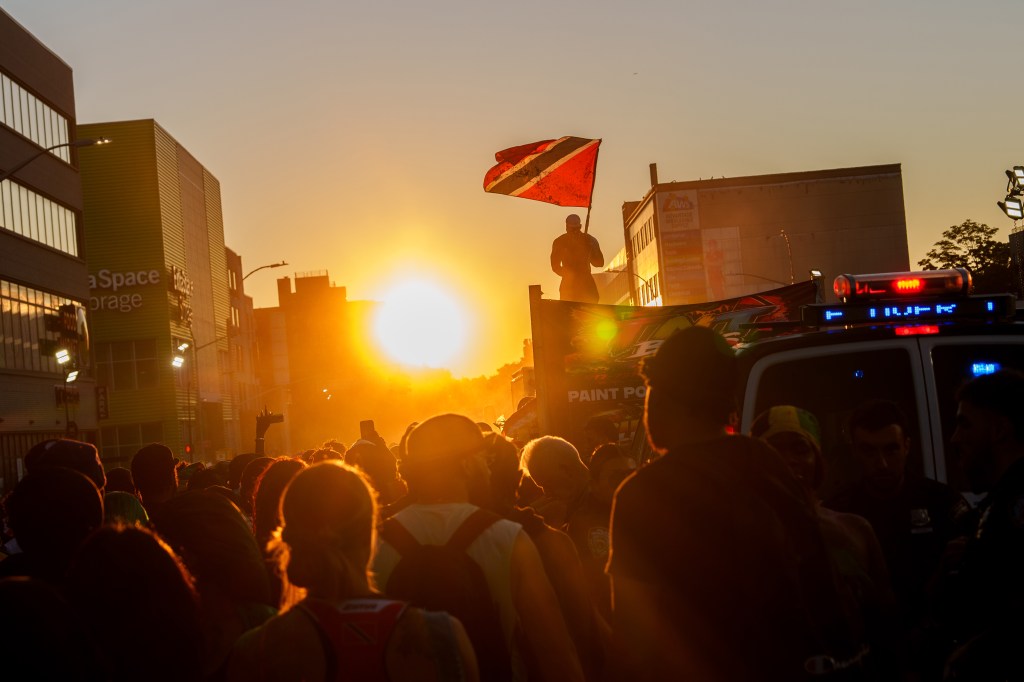 A crowd of people at the J'ouvert celebration at sunrise in Brooklyn, with a person on a truck holding a Trinidad flag.