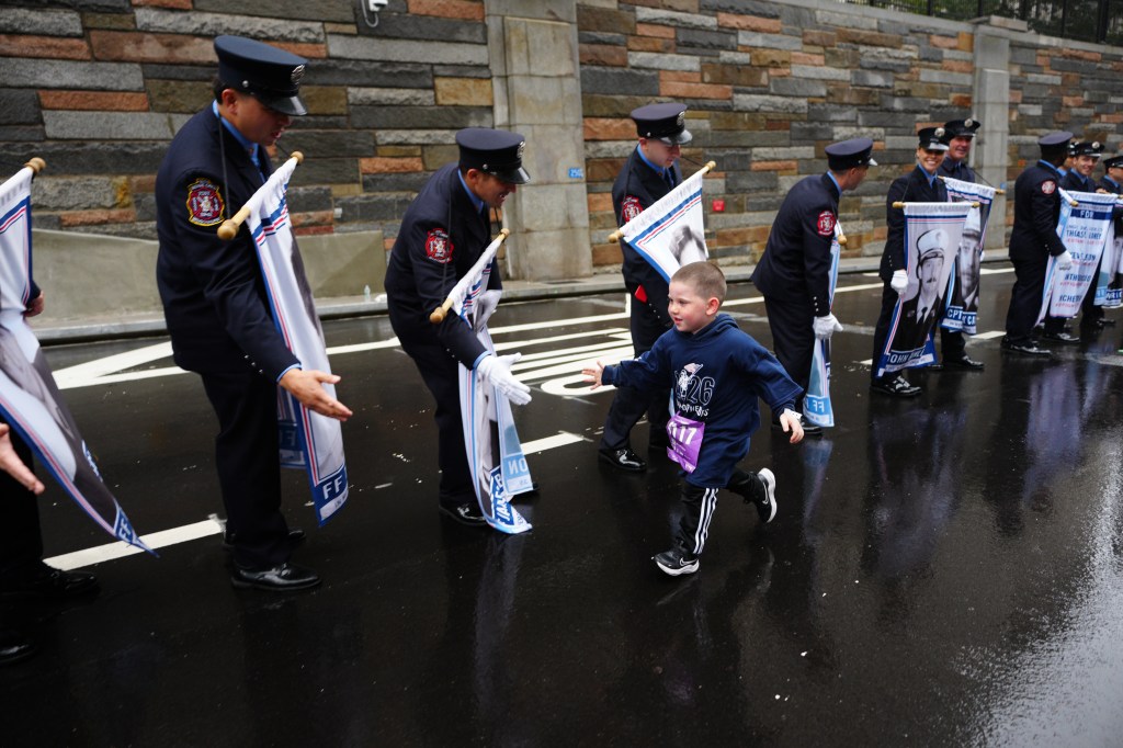 A young boy with a running bib participates in the 25th annual Tunnel to Towers NYC 5K Run & Walk, surrounded by firefighters holding banners.