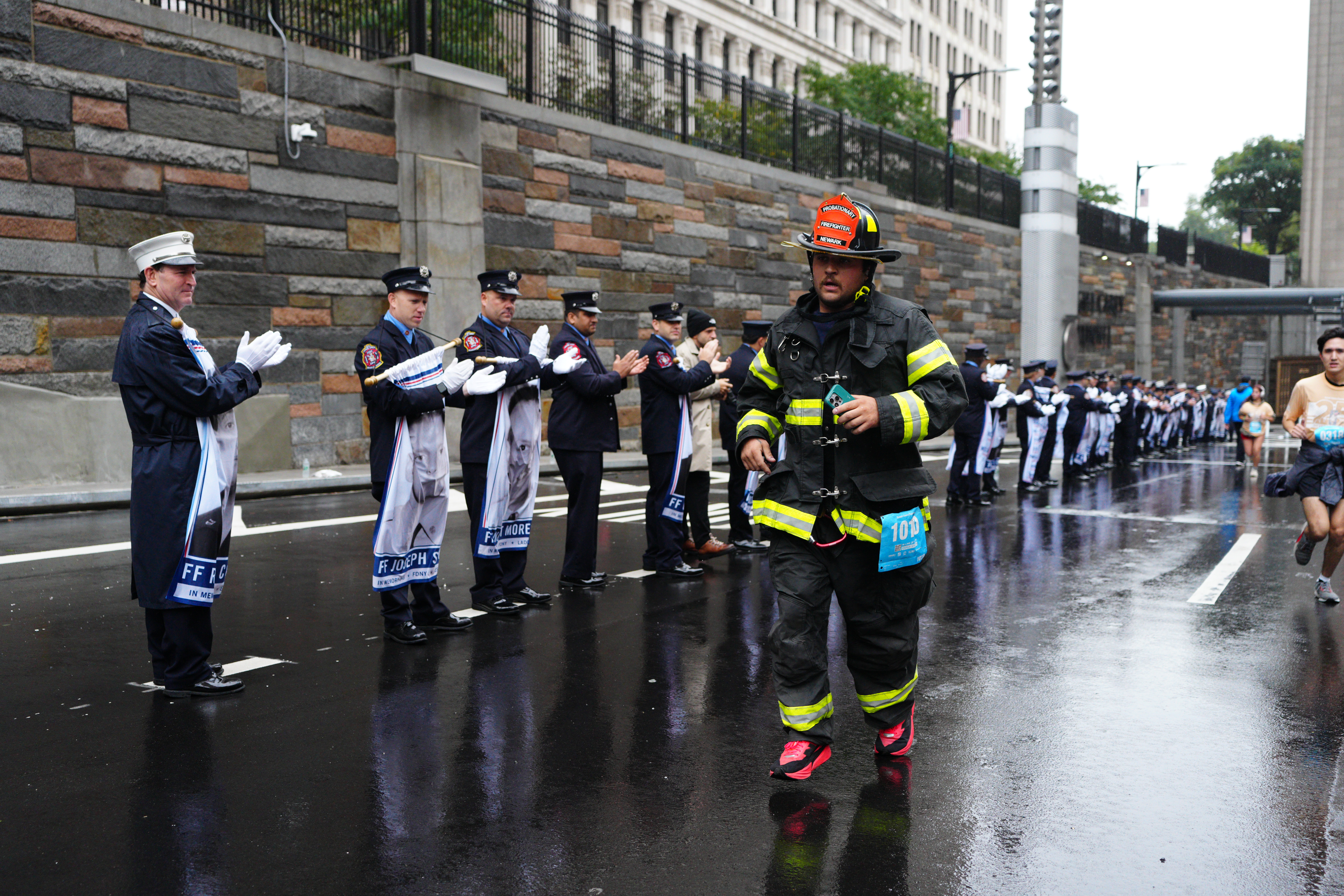 A runner in full firefighter gear passes uniformed firefighters clapping along the race route of the Tunnel to Towers 5K.