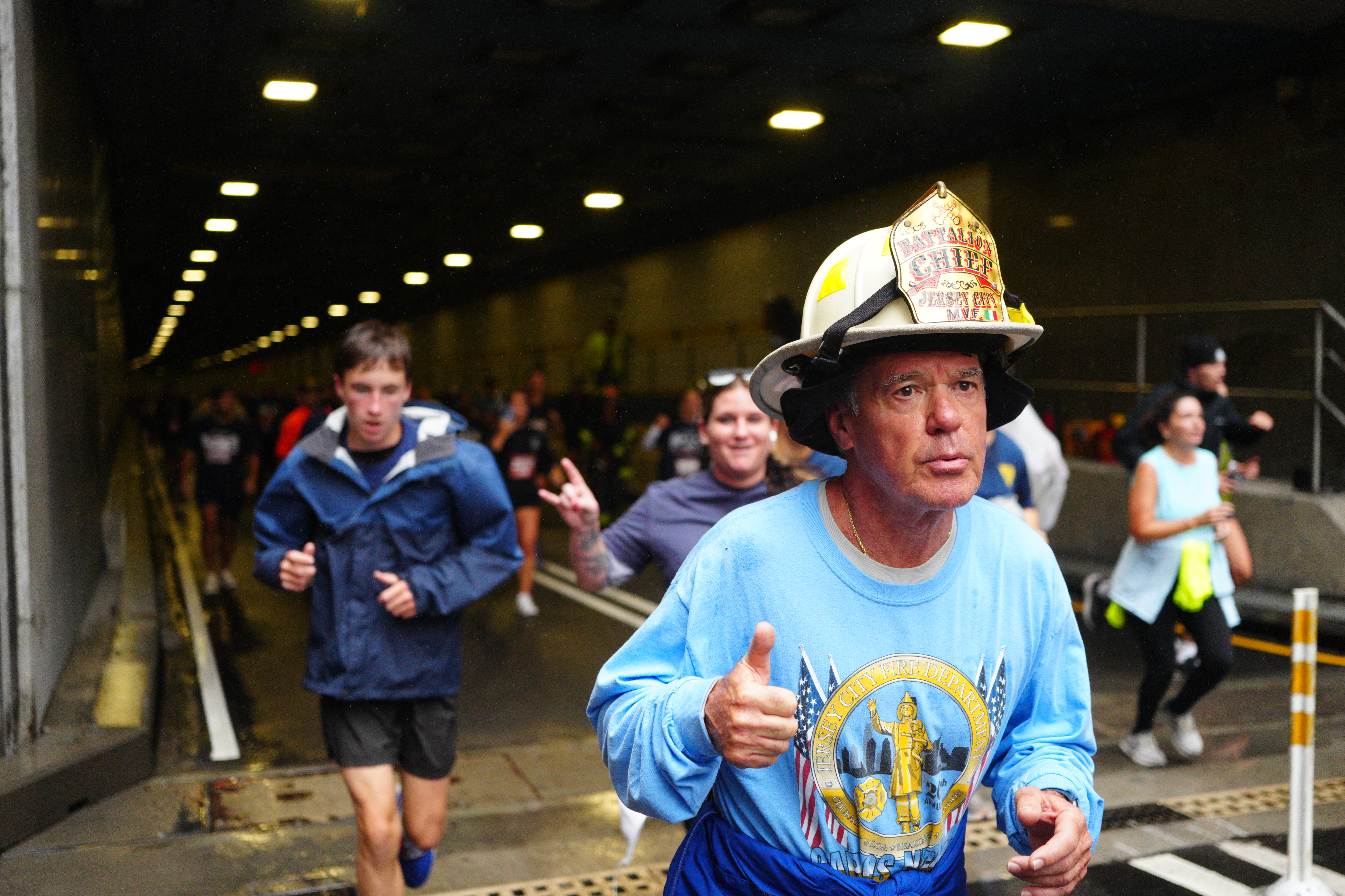 A man in a light blue shirt, giving a thumbs up, and wearing a "Battalion Chief Jersey City" firefighter helmet, participates in the 25th annual Tunnel to Towers NYC 5K Run & Walk.