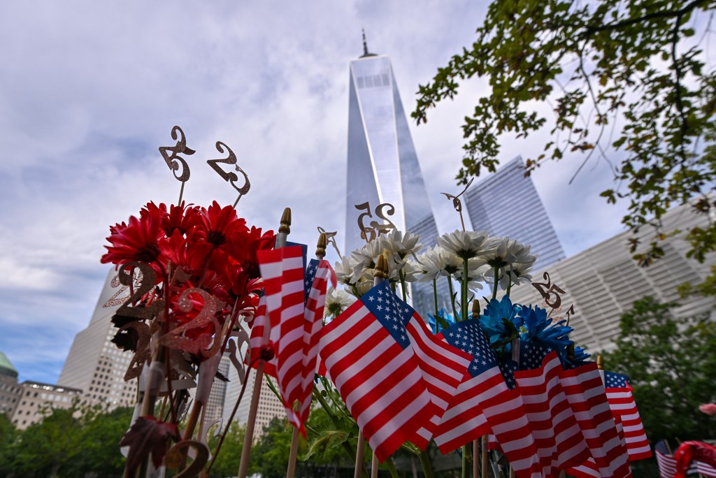 Small American flags, red and white flowers, and "25" markers sit in the foreground of the One World Trade Center and other buildings in New York City.