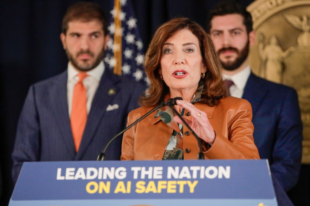 New York Governor Kathy Hochul speaking at a press conference on AI safety, flanked by two men in suits.