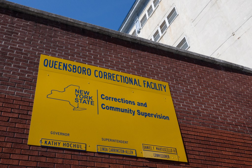 A yellow sign for Queensboro Correctional Facility on a brick wall.