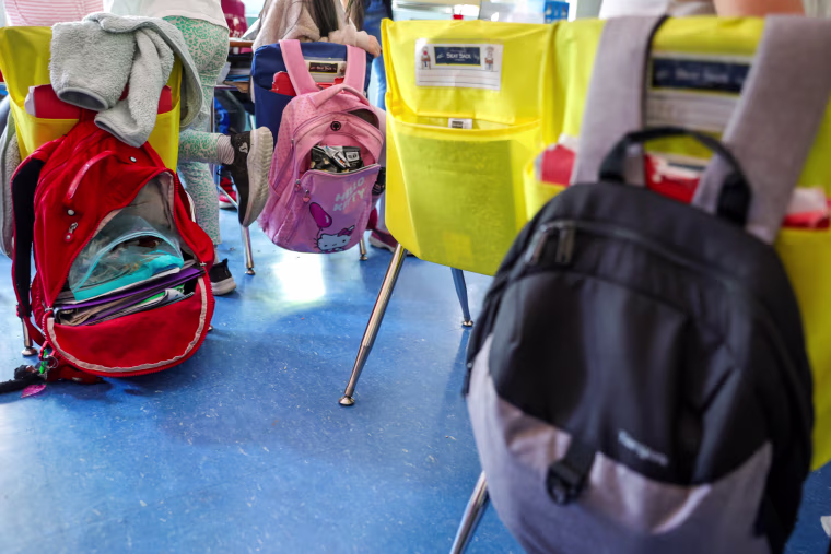 Student backpacks hang on the backs of classroom chairs.