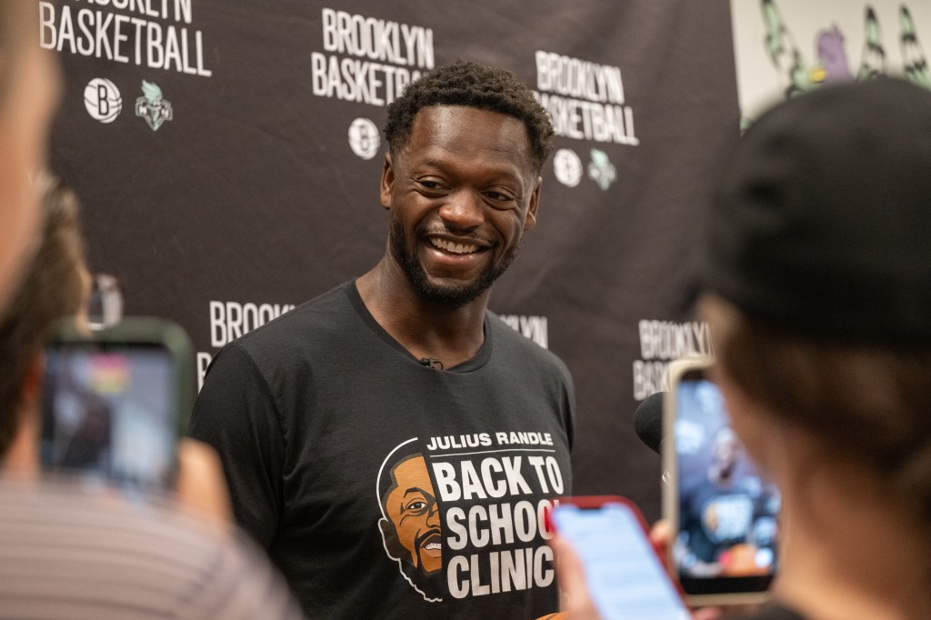 Julius Randle smiling while wearing a "Back to School Clinic" t-shirt and being filmed by members of the media.