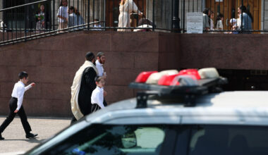 A police car is parked in front of a building as men and boys in traditional Jewish attire walk nearby. Other people are visible on the upper level behind a railing.