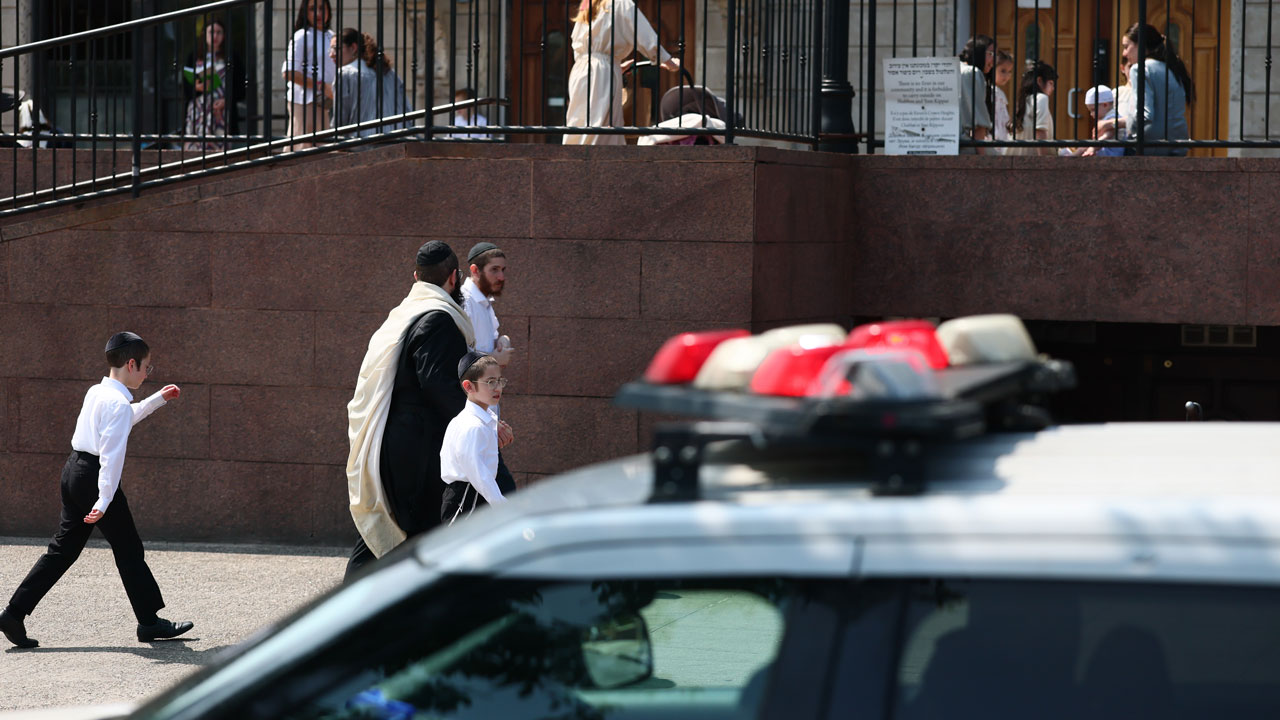 A police car is parked in front of a building as men and boys in traditional Jewish attire walk nearby. Other people are visible on the upper level behind a railing.