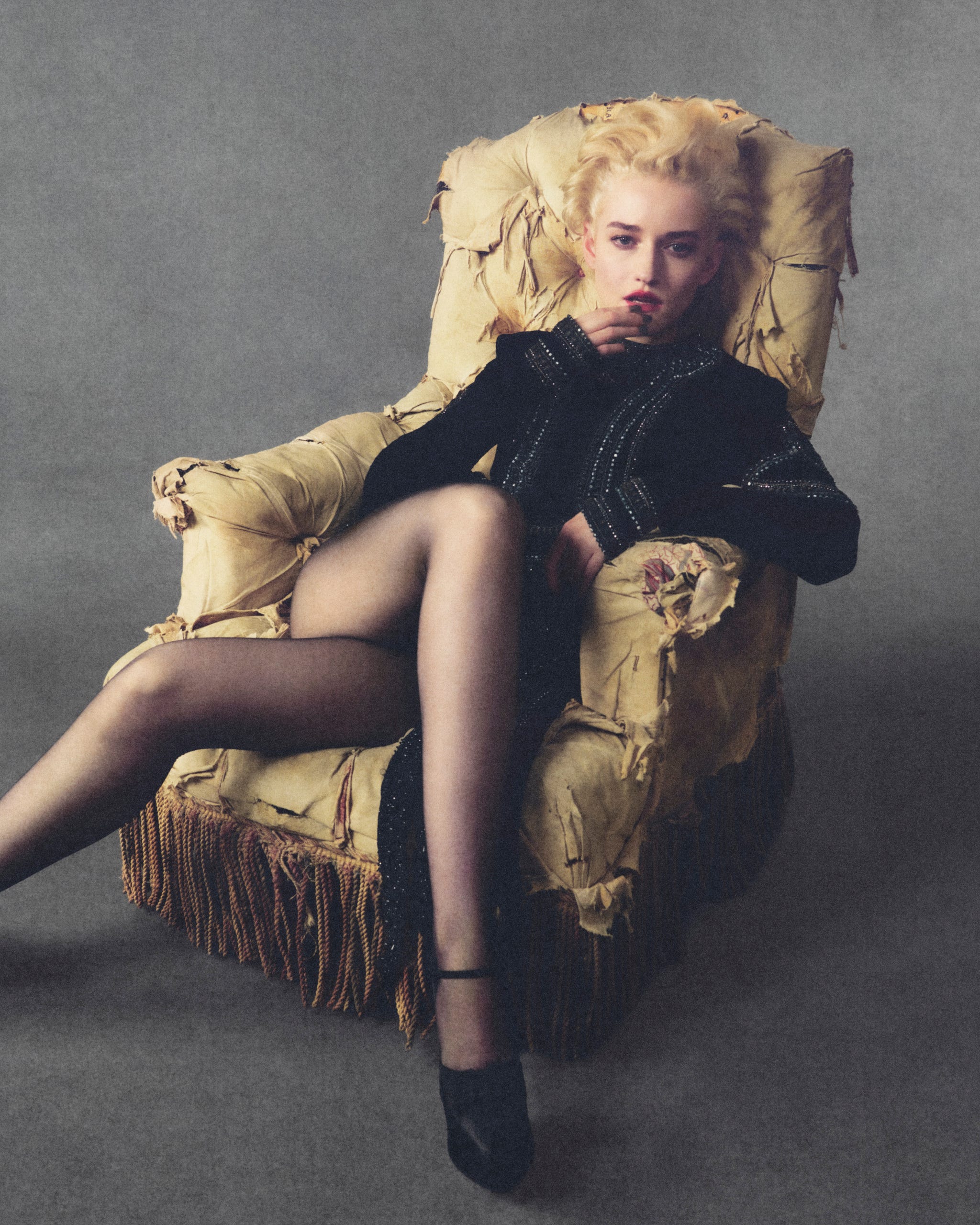 Julia Garner poses confidently in a vintage armchair, wearing a stylish black outfit and heels.