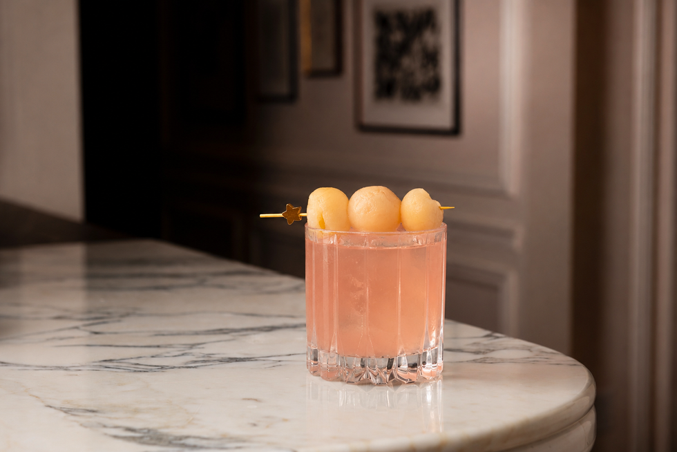 A pink cocktail garnished with melon balls in a textured glass on a marble table.