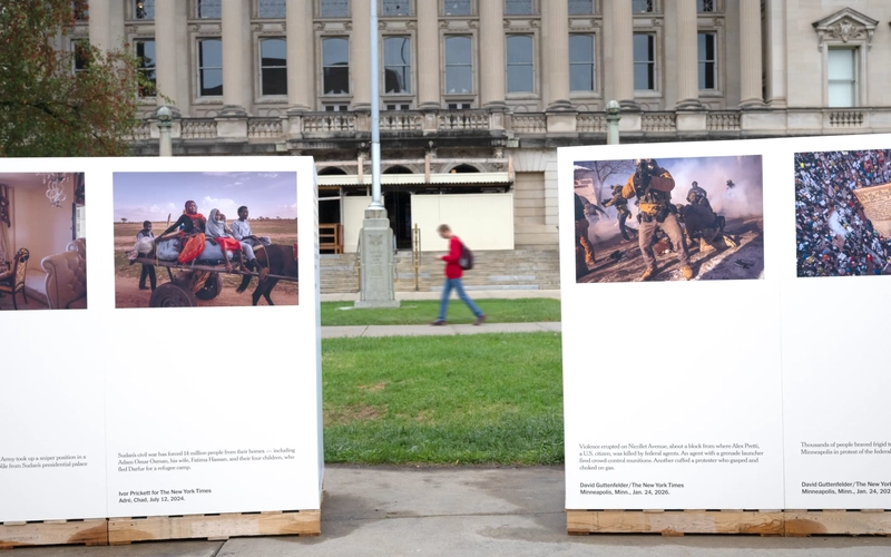 An outdoor photo exhibit is set up outside, with images displayed on structures mounted on wooden shipping pallets. In the background across a grassy lawn, a person in a red shirt walks past a large classical stone building with prominent columns.
