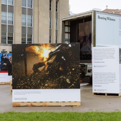 An outdoor pop-up exhibit display titled Bearing Witness features photojournalism prints set up on wooden pallets against a stone building backdrop.
