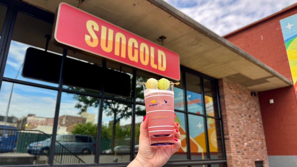 A hand holding a Honey Deuce cocktail in front of a storefront sign that reads "SUNGOLD.