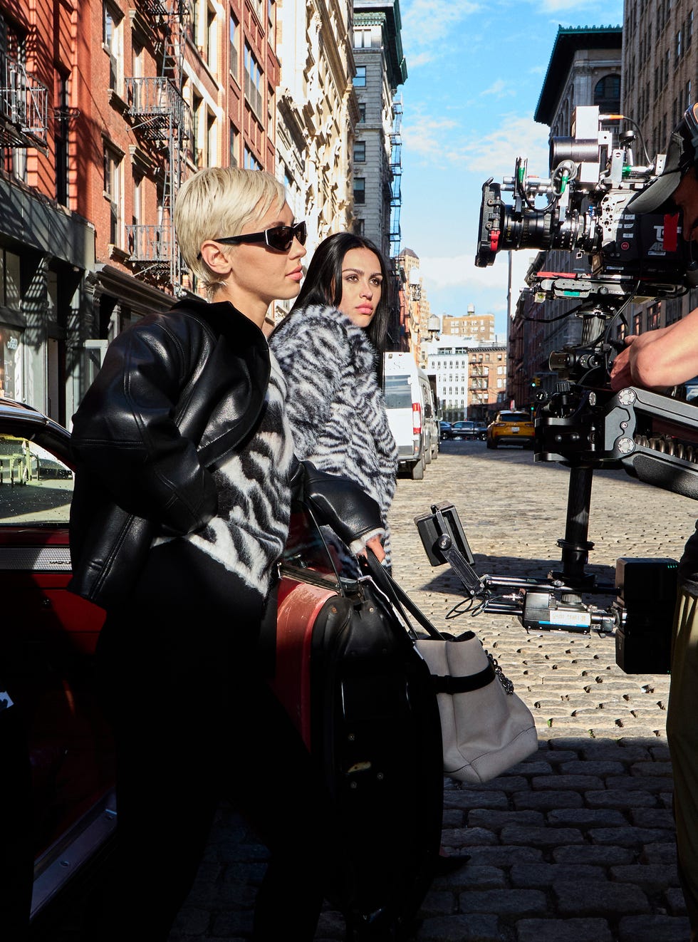 Two stylish women stand in a city street, one holding a bag while a camera crew is filming nearby.