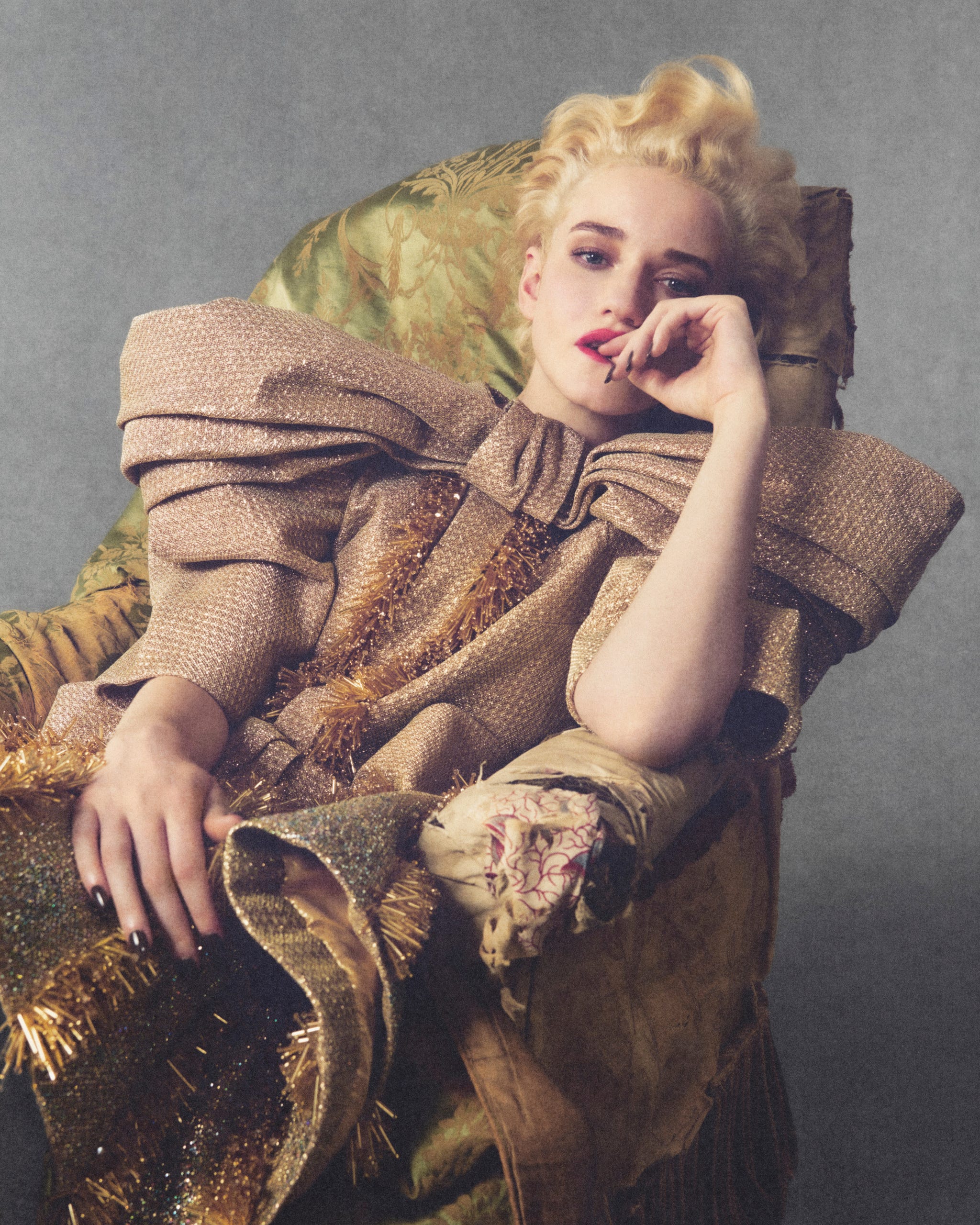 Julia Garner in an extravagant gold outfit reclines thoughtfully on an ornate green chair.
