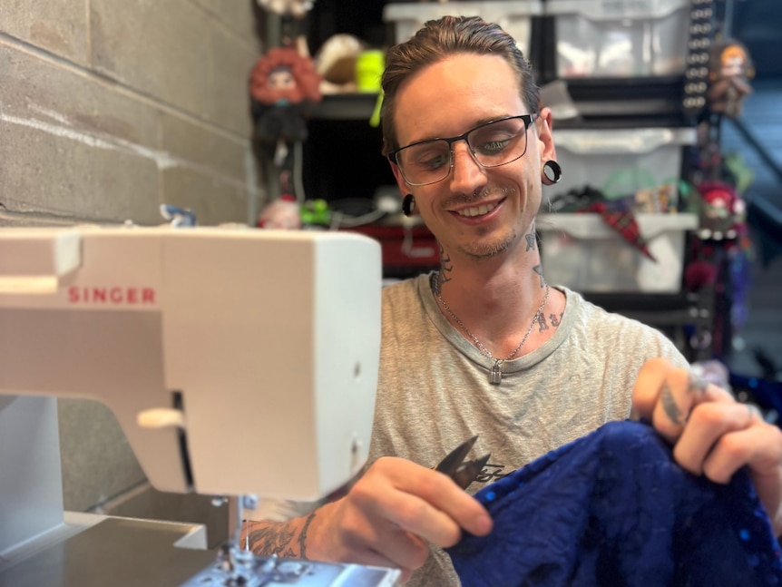 A smiling person holds fabric and scissors while sat in front of a sewing machine.