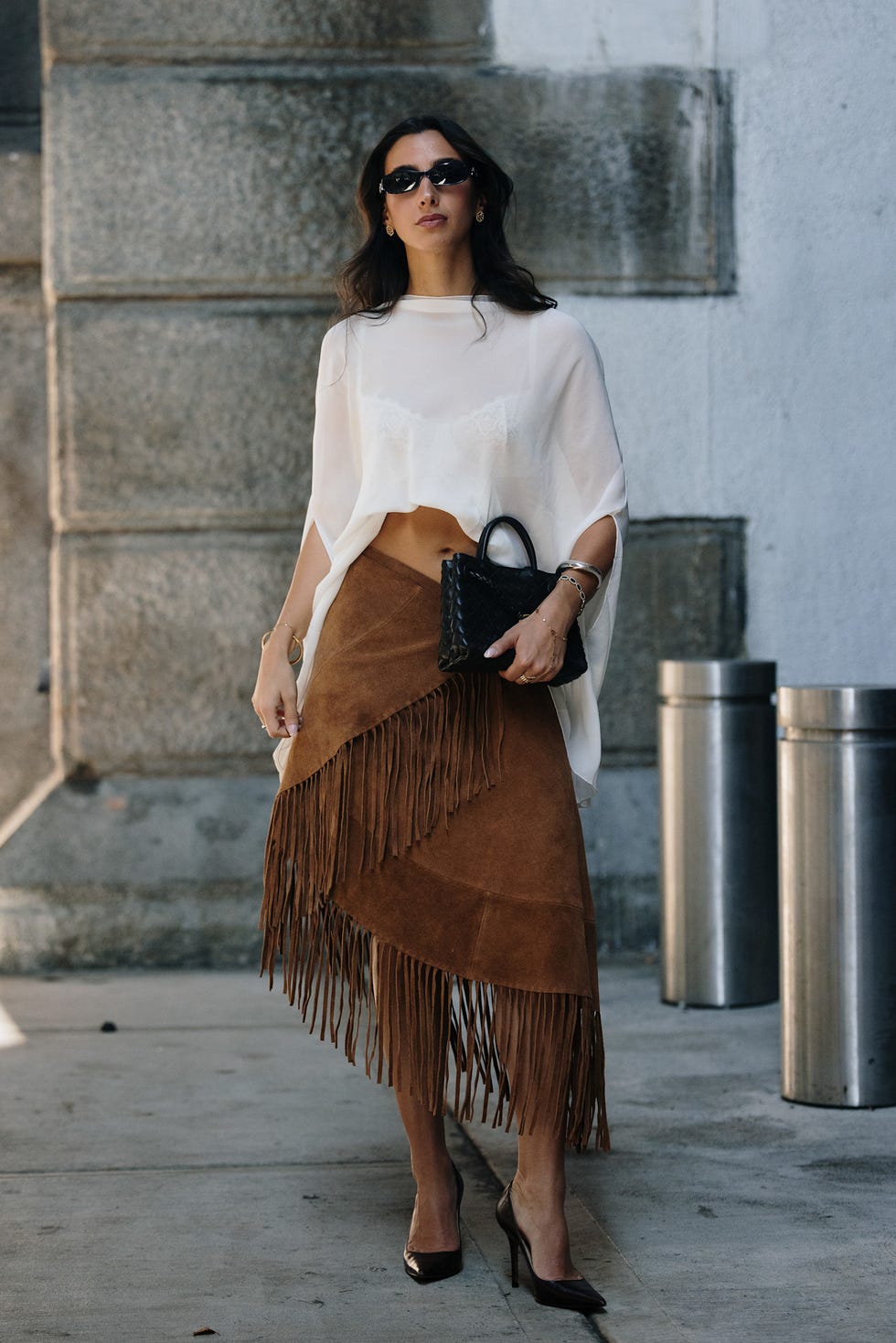 Woman in a sheer white top and fringed brown skirt, wearing sunglasses and holding a small black handbag. Woman in a sheer white top and fringed brown skirt, wearing sunglasses and holding a small black handbag.