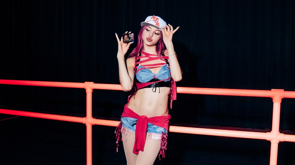 A woman with pink hair wearing a jean bustier and white baseball cap poses with a pack of 5 Gum.