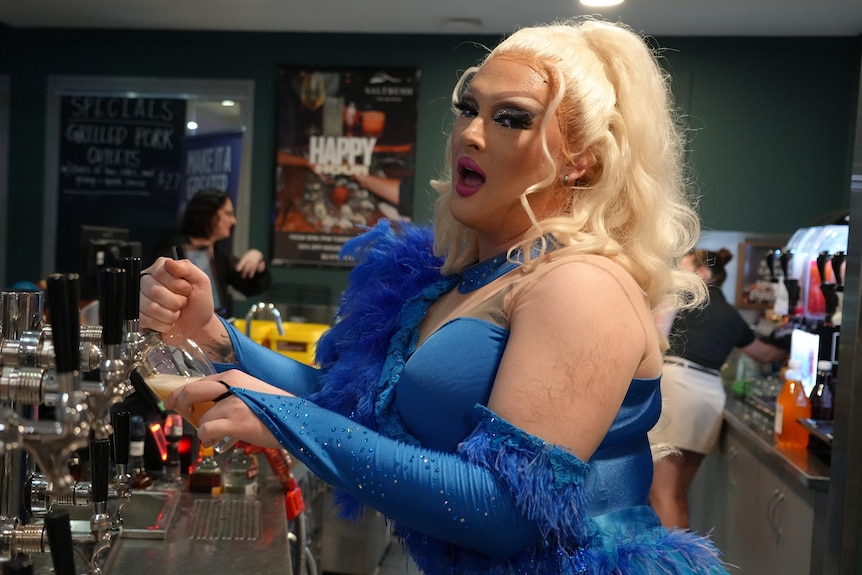 A drag queen in a blonde wig and blue dress pours a tap beer behind a bar.
