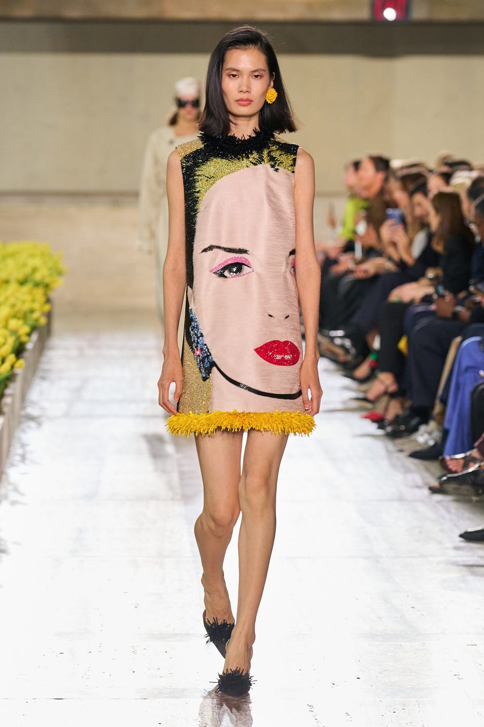 Model walking on runway wearing a dress featuring a colorful face design with fringe details.