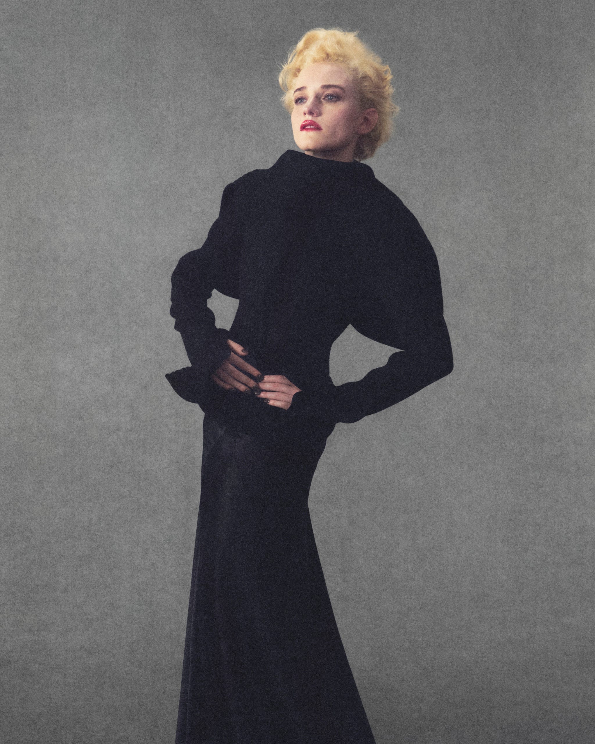Julia Garner in a dramatic black gown poses confidently against a muted gray background.