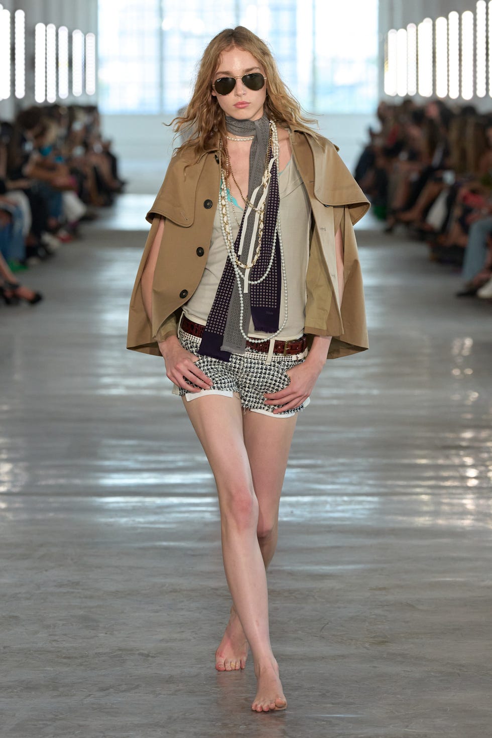 Model walking down the runway wearing stylish sunglasses and layered clothing in a fashion show setting.