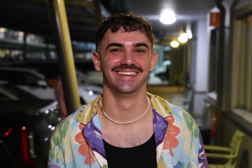 A young man with a moustache and brunette hair smiles in a dimly lit car park, wearing a colourful buttoned shirt.
