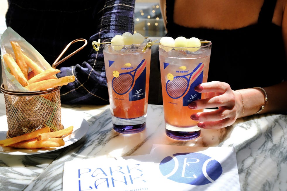 Two cocktails garnished with fruit next to a basket of fries on a marble table, with a menu visible.