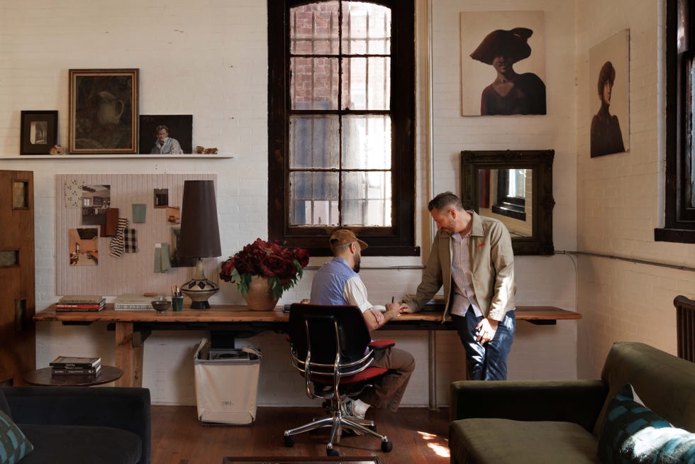 Two men discussing at a desk in a stylish, sunlit room with artwork on the walls and flowers on the table.