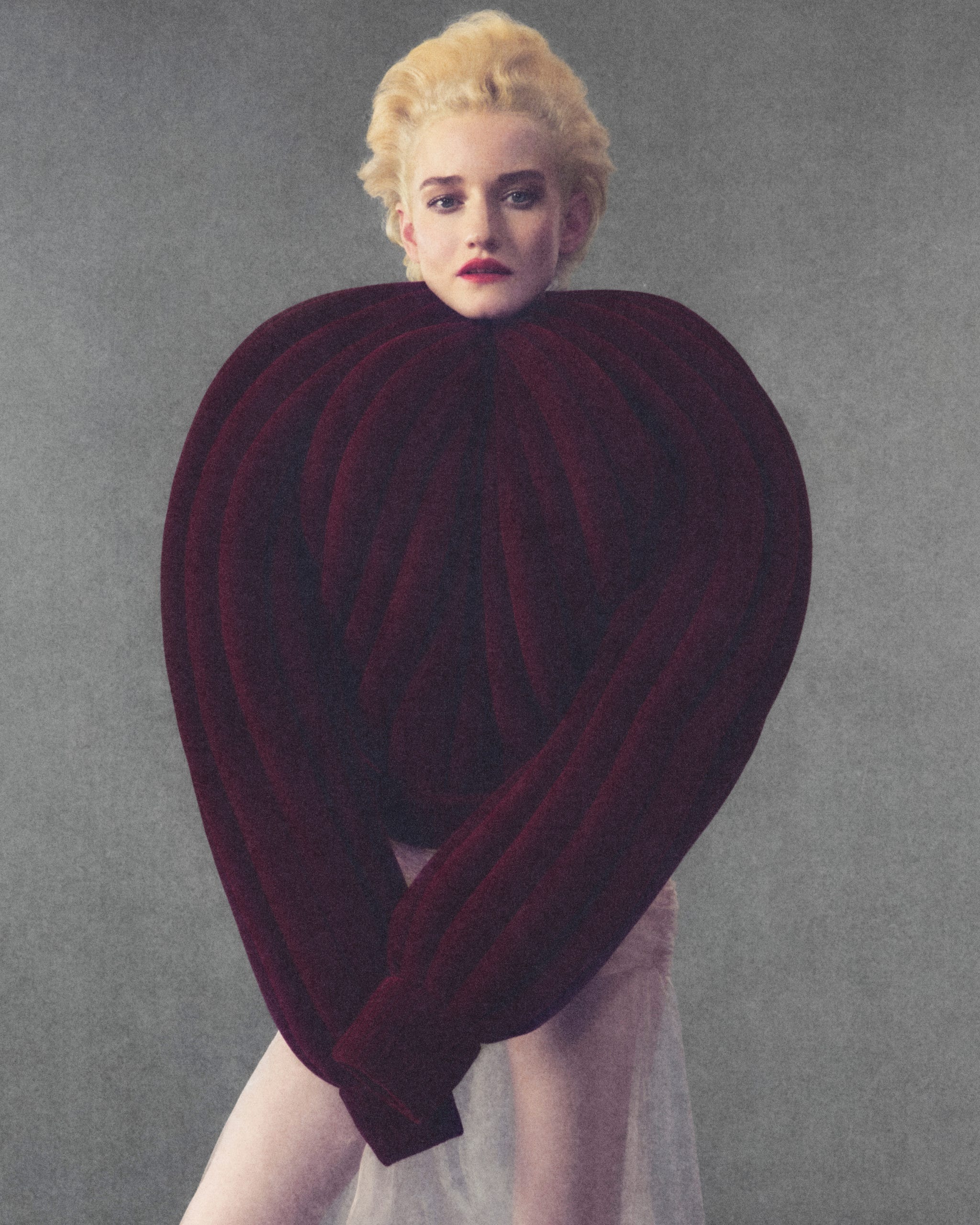 Julia Garner wearing a large, burgundy, heart-shaped garment with an artistic design set against a neutral background.