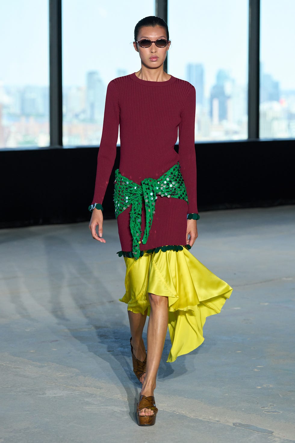 Model walking on runway wearing a burgundy dress with a green sash and a yellow skirt, accessorized with sunglasses.