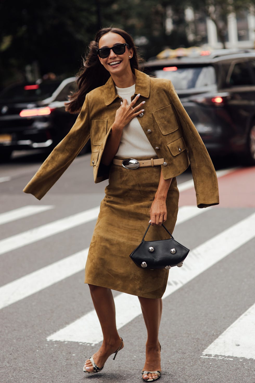 Woman in a stylish brown suede outfit smiling while walking across a crosswalk, holding a small black handbag. Woman in a stylish brown suede outfit smiling while walking across a crosswalk, holding a small black handbag.