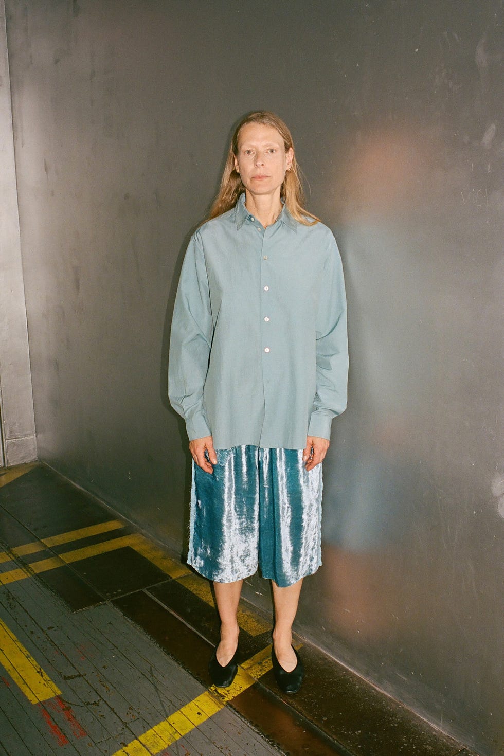 A person wearing a light green shirt and silver skirt stands against a gray wall in a minimalist setting.