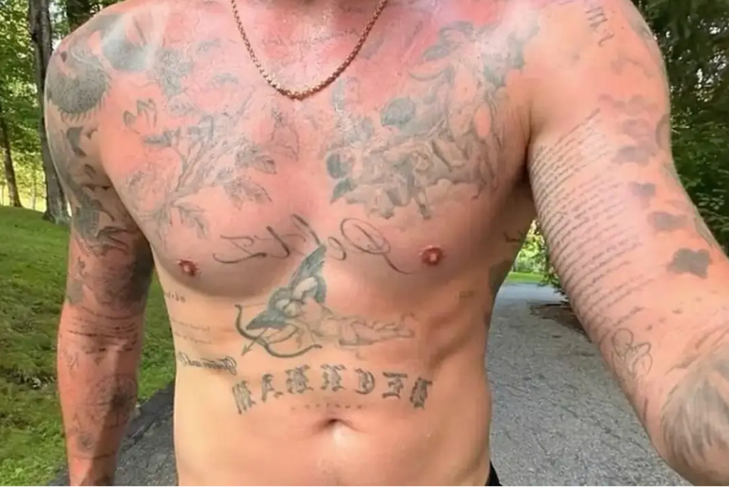 A person with extensive tattoos, a necklace, and no shirt stands on a path.