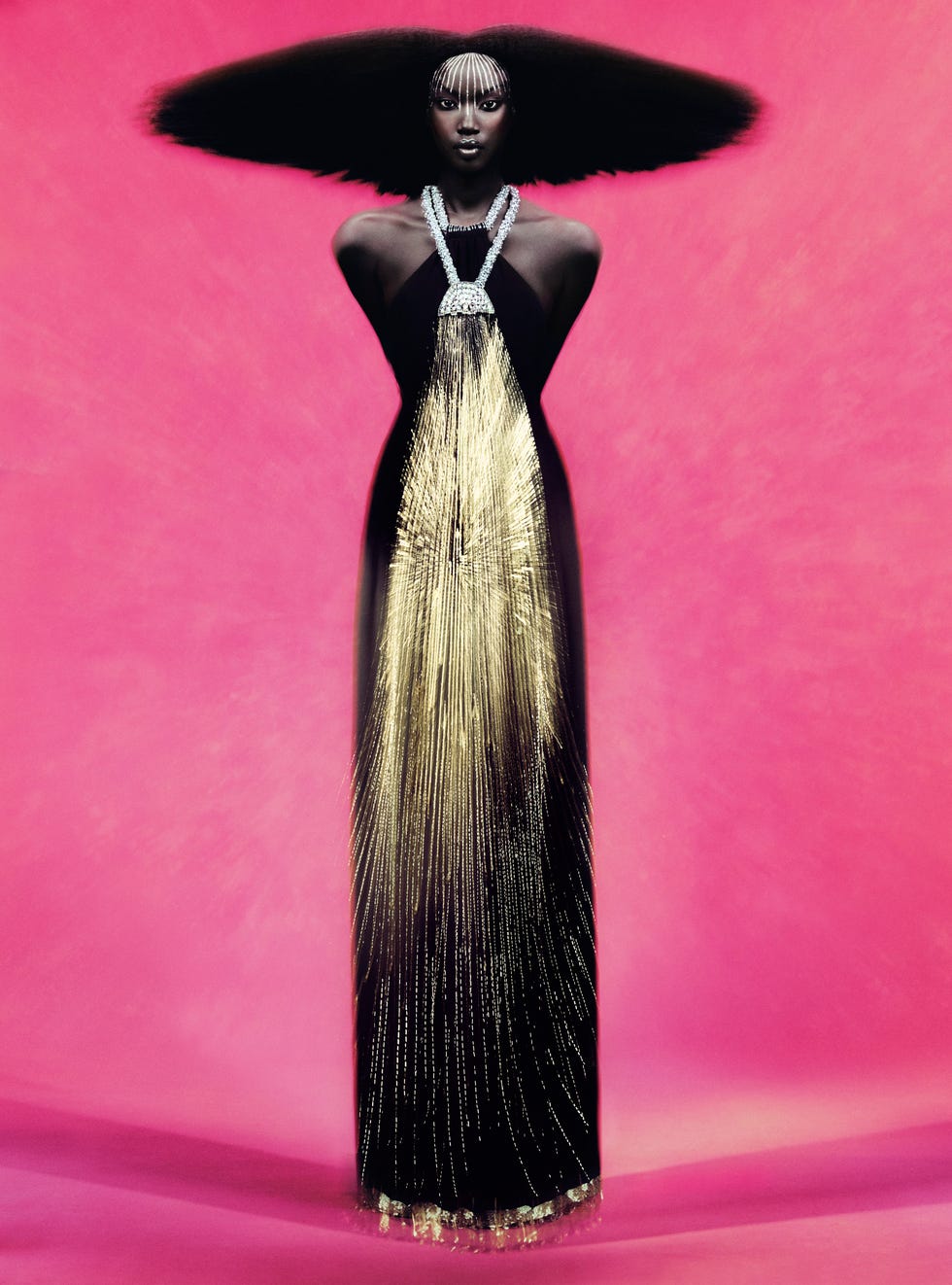 A striking fashion model in a dramatic black and gold dress against a pink backdrop.