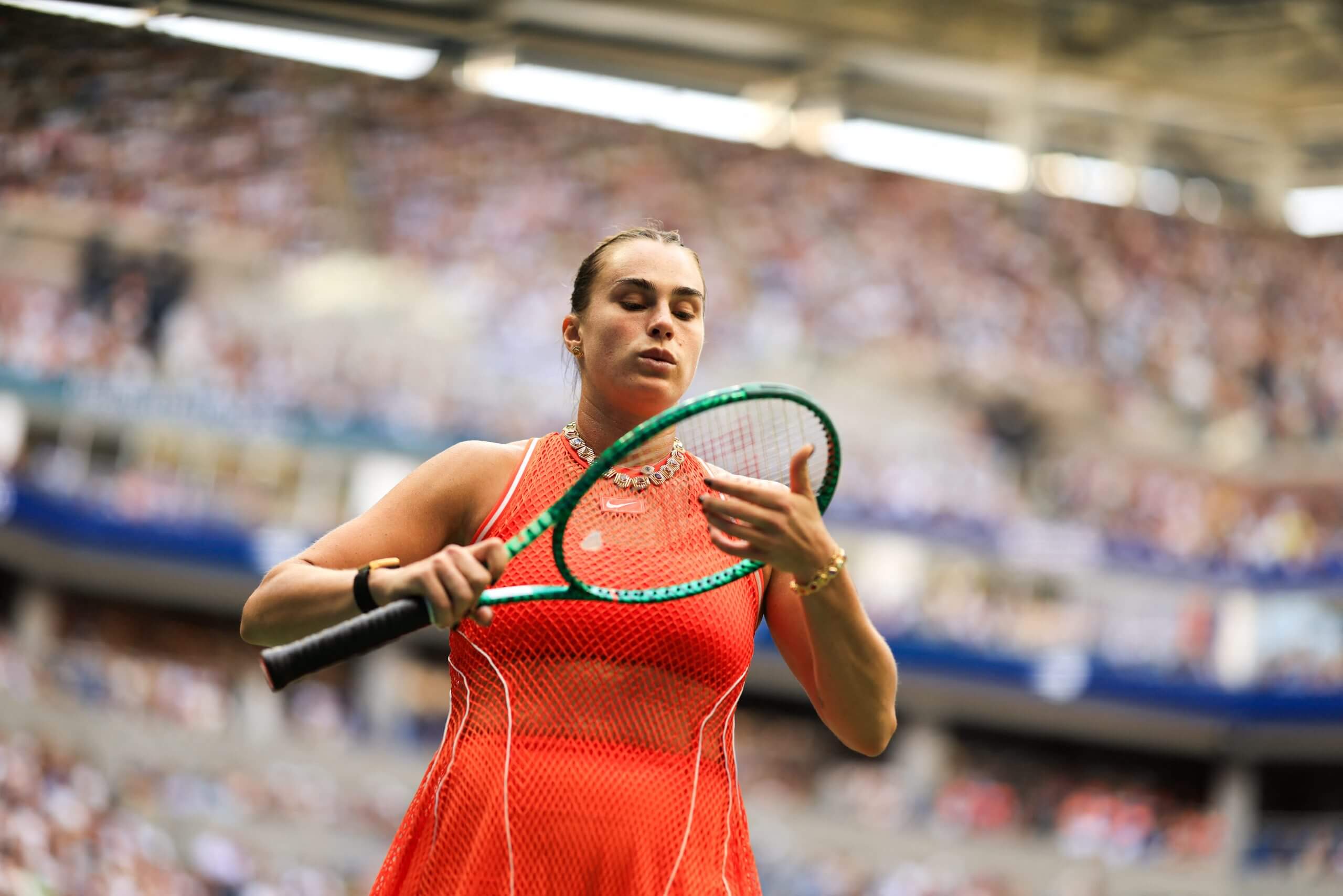 Aryna Sabalenka adjusts her strings with fans all around her in soft focus inside a tennis stadium.