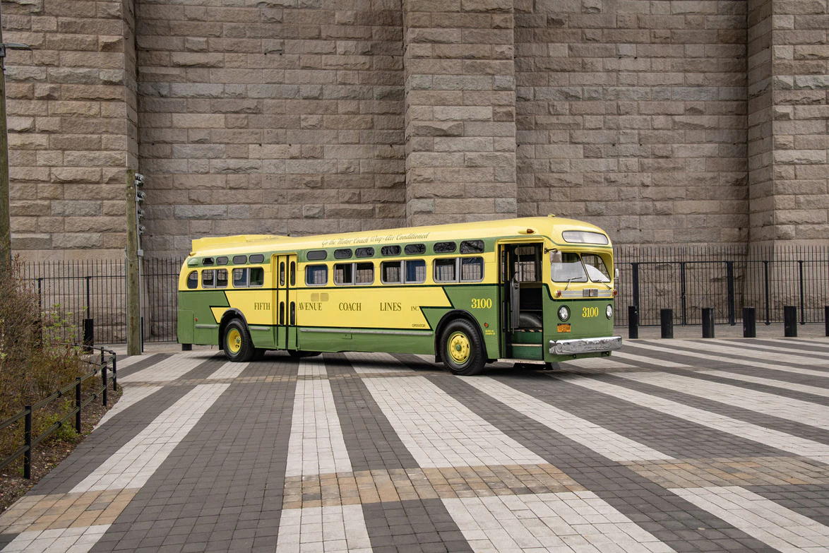 Vintage green and yellow bus parked on a patterned stone plaza with a large stone wall in the background.