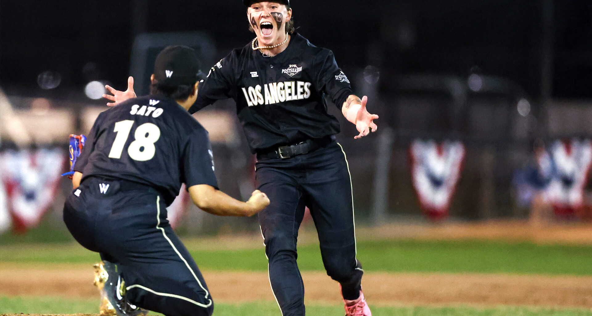 Two baseball players in black uniforms celebrate on the field, one running toward a kneeling teammate with her arms outstretched.