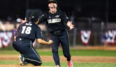 Two baseball players in black uniforms celebrate on the field, one running toward a kneeling teammate with her arms outstretched.
