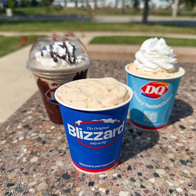 Three Dairy Queen desserts sit on a speckled outdoor table: two Blizzards in blue cups with whipped cream and a chocolate sundae in a clear cup with chocolate drizzle.