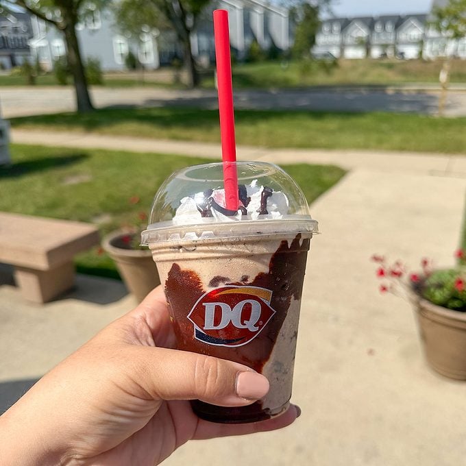 A hand holds a Dairy Queen cup filled with a chocolate sundae topped with whipped cream and chocolate drizzle, outside on a sunny day.