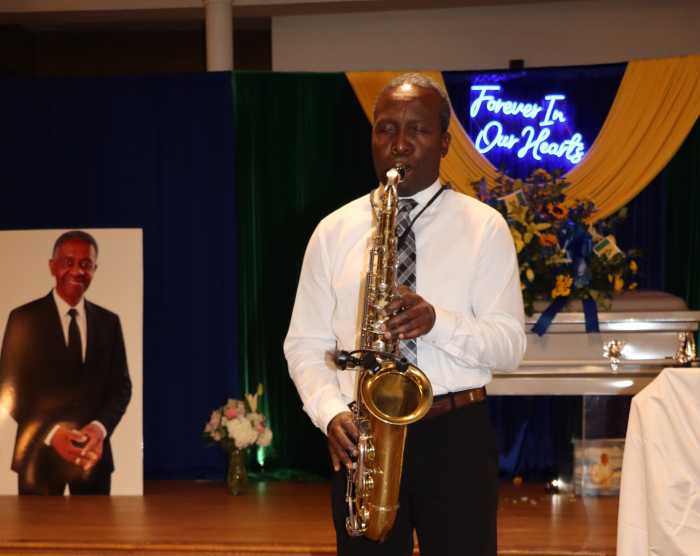 Vincentian calypsonian and musician Ulric Hamlett plays the saxophone, with a photo of Granville Straker in background.